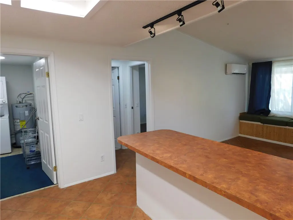A bright room with white walls, brown tile floors, and a long brown countertop. A laundry room and hallway are visible through open doorways.