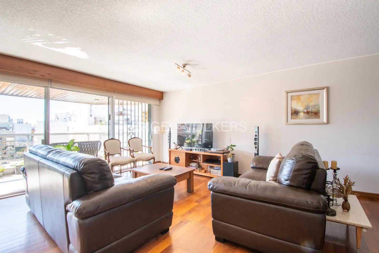 Bright living room with hardwood floors, brown leather sofas, and a balcony view of the city.