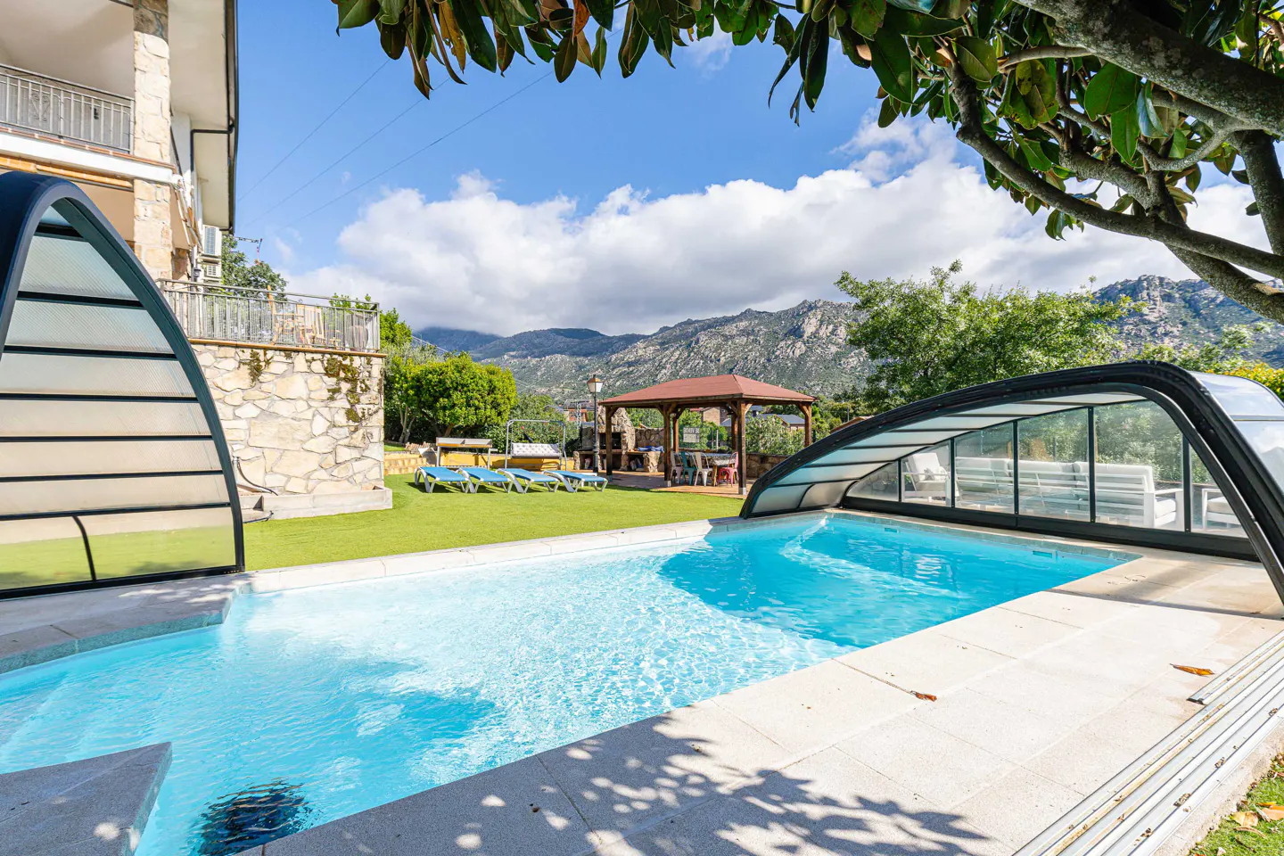 Outdoor pool with a retractable cover, surrounded by green grass, lounge chairs, and a gazebo, with mountains in the background.