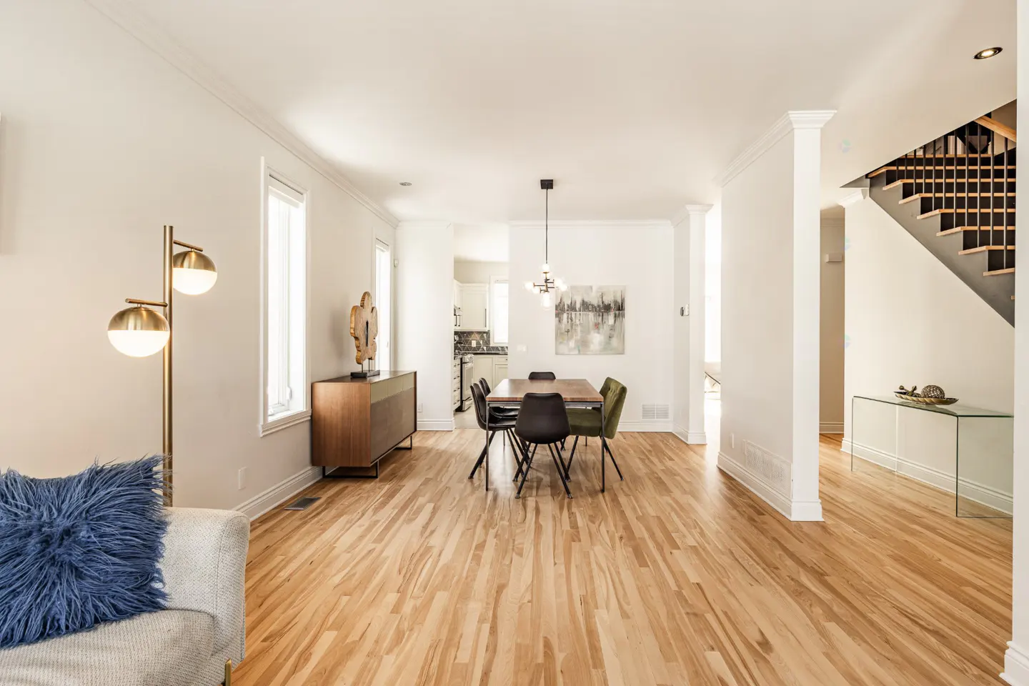 Bright, open-concept living and dining area with hardwood floors, white walls, and modern furnishings. Staircase visible in the background.