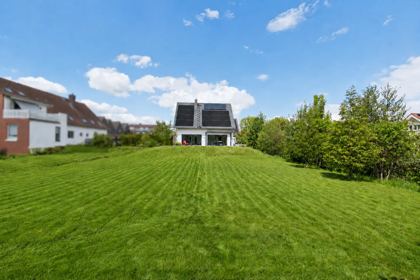 A modern white house with solar panels sits atop a grassy hill under a blue, cloudy sky.