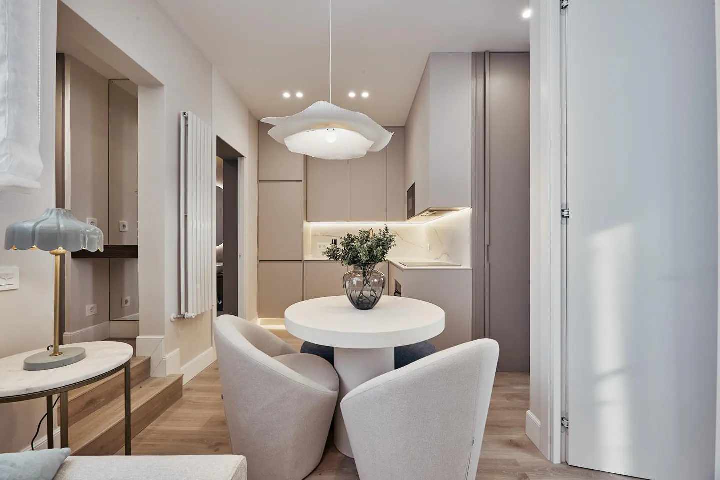 A modern apartment interior with a round white table, three chairs, and a kitchen in the background.