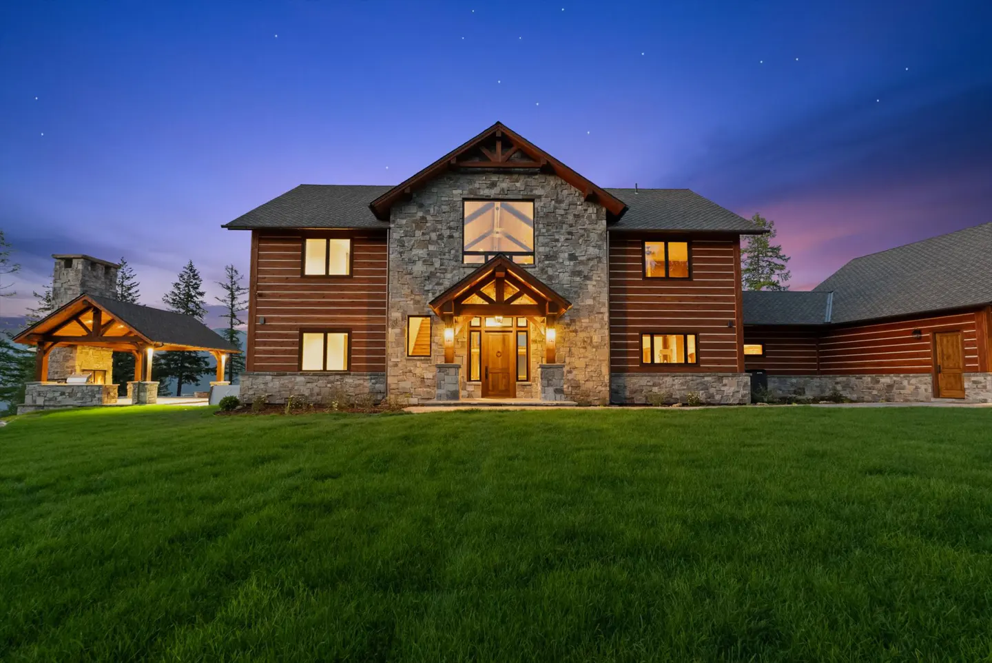Two-story log cabin home with stone accents, a covered patio with fireplace, and a green lawn under a twilight sky.