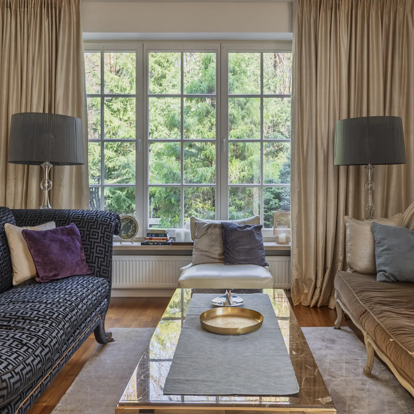 Living room with a blue patterned sofa, brown chaise lounge, and a marble coffee table. Large window with beige curtains overlooks green trees.