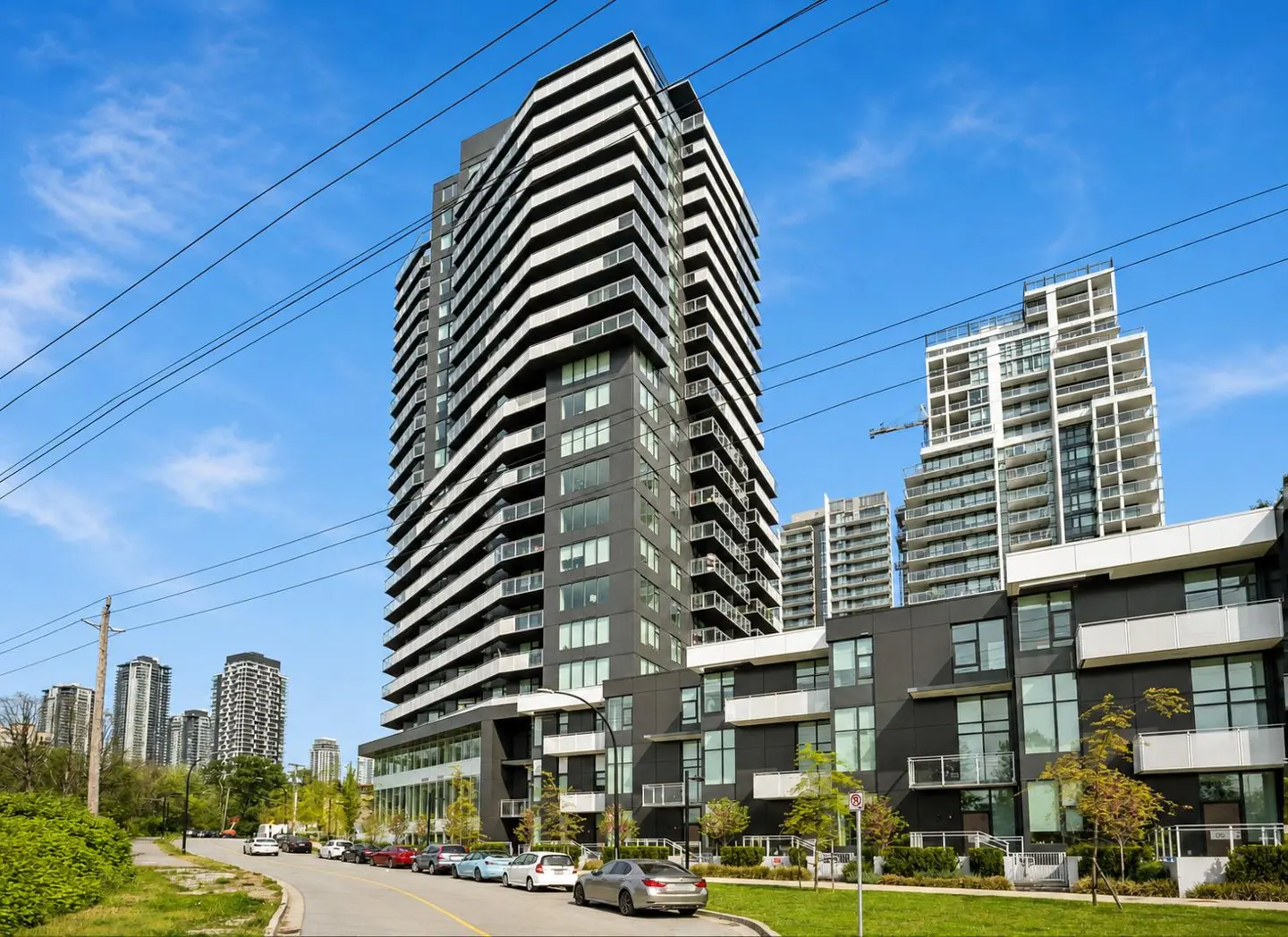 Modern black and white high-rise condo building with balconies under a blue sky. Cars line the street in front of the building.