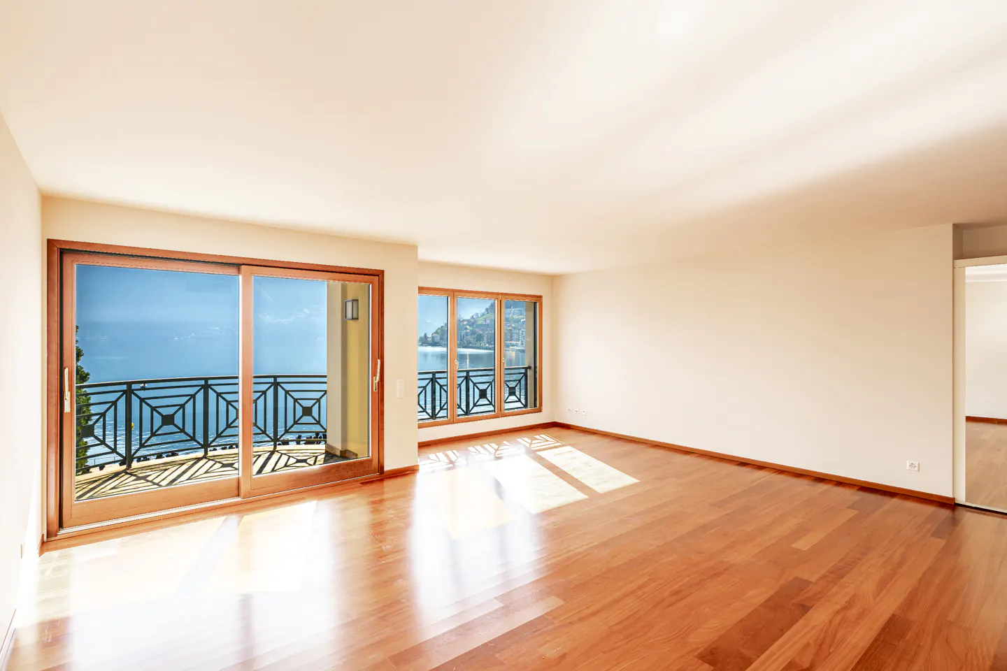 Bright, empty room with hardwood floors and white walls. Sliding glass doors open to a balcony with a view of a blue lake.