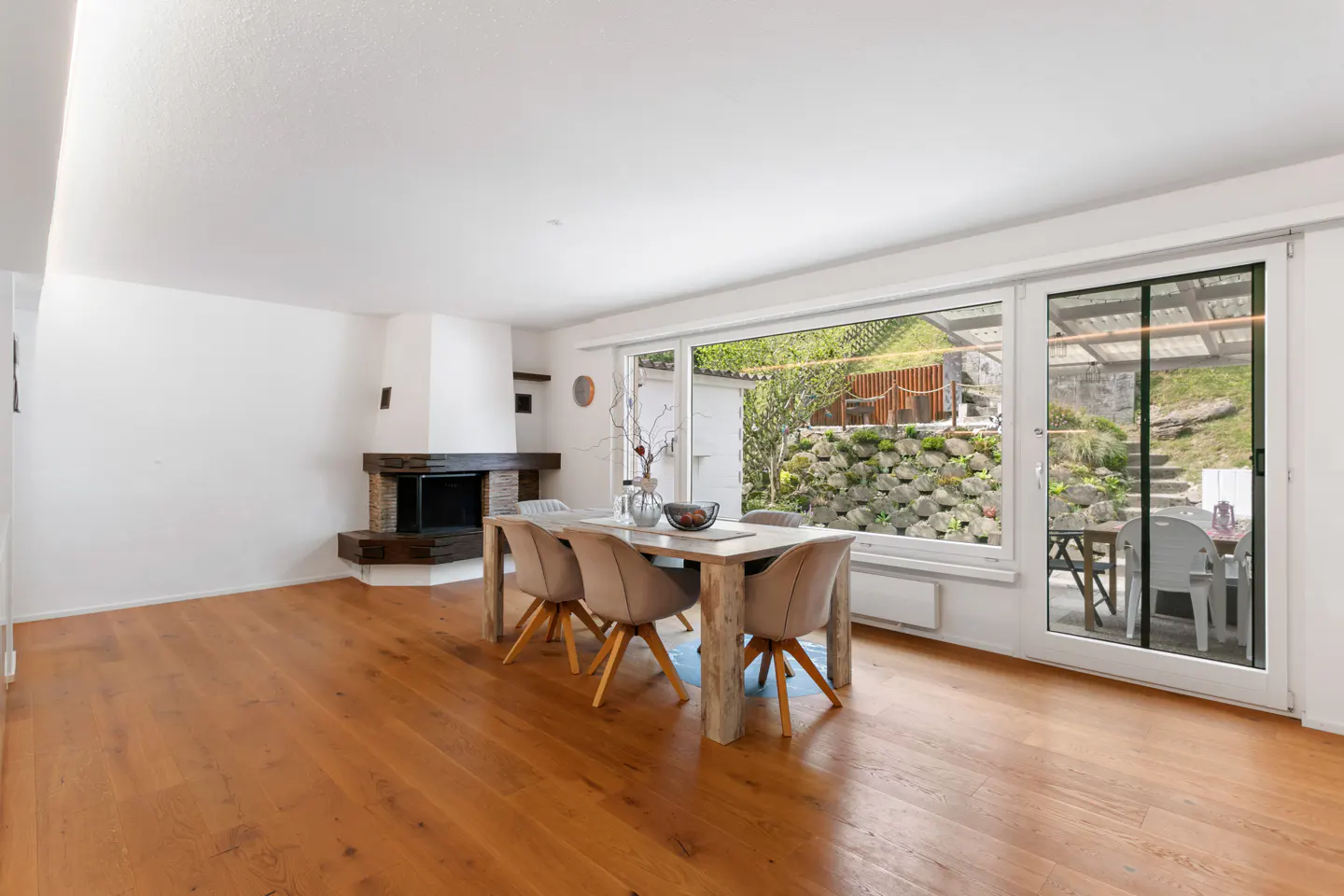 Bright dining room with wood floors, a stone fireplace, and a large window overlooking a terraced garden. A wooden table with six chairs sits in the center.
