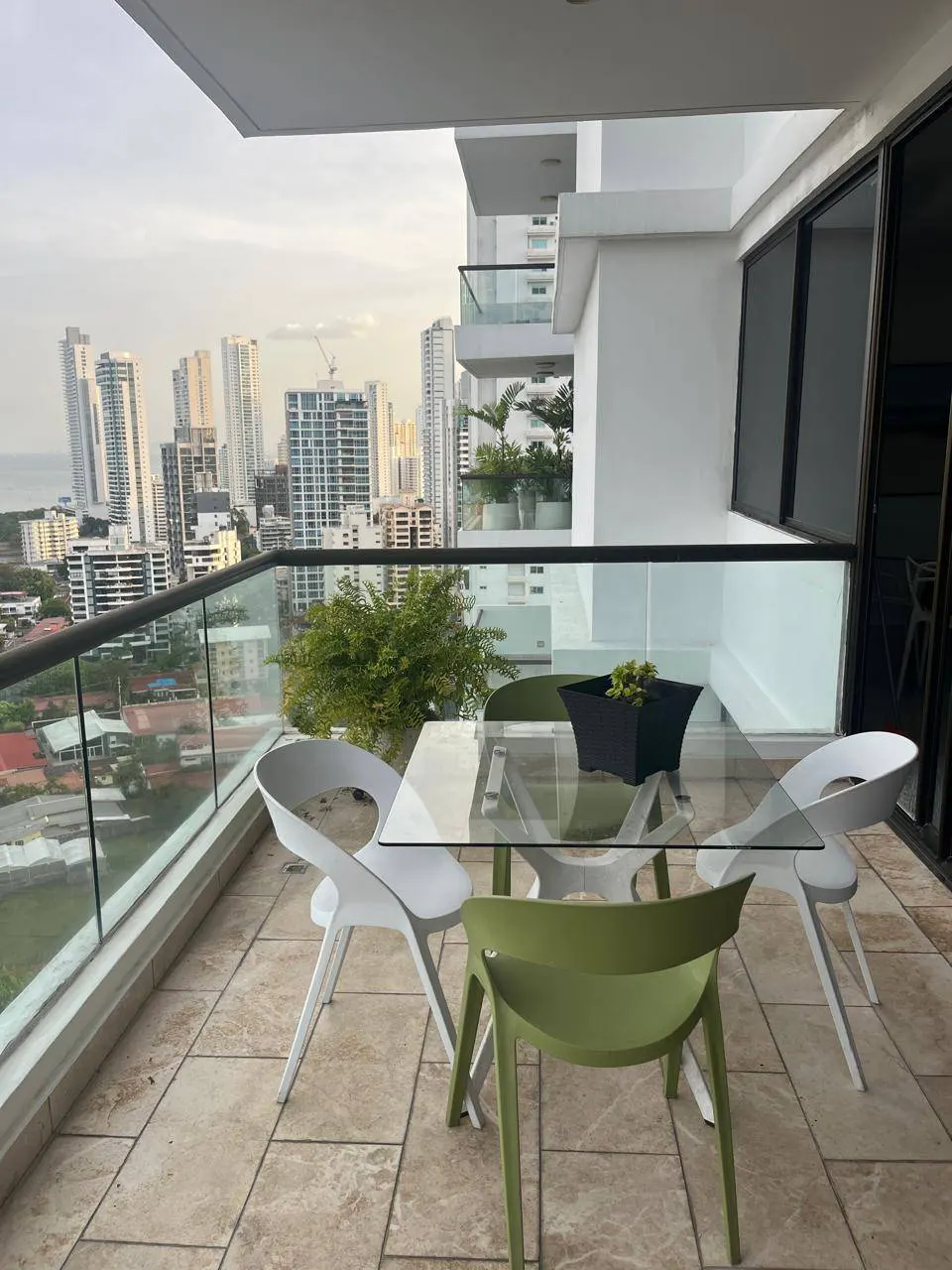 Balcony with glass table, white and green chairs, and a city view. A black planter sits on the table.