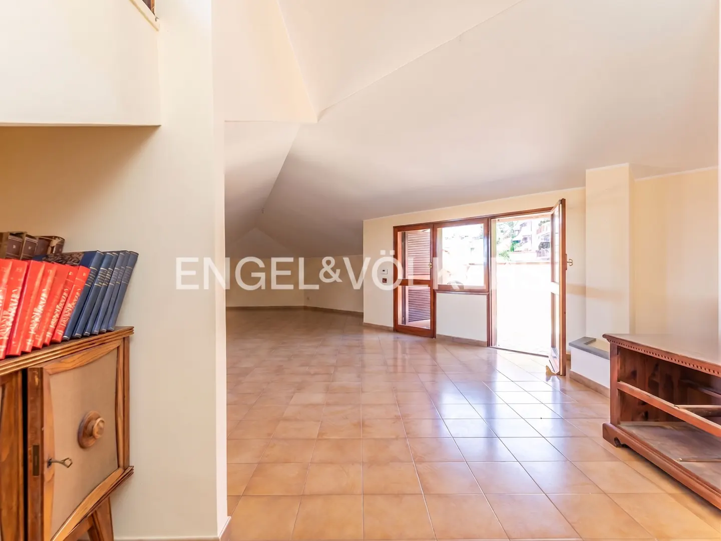 Spacious attic room with light tile floors, sloped white ceilings, and an open door leading to a bright outdoor terrace.
