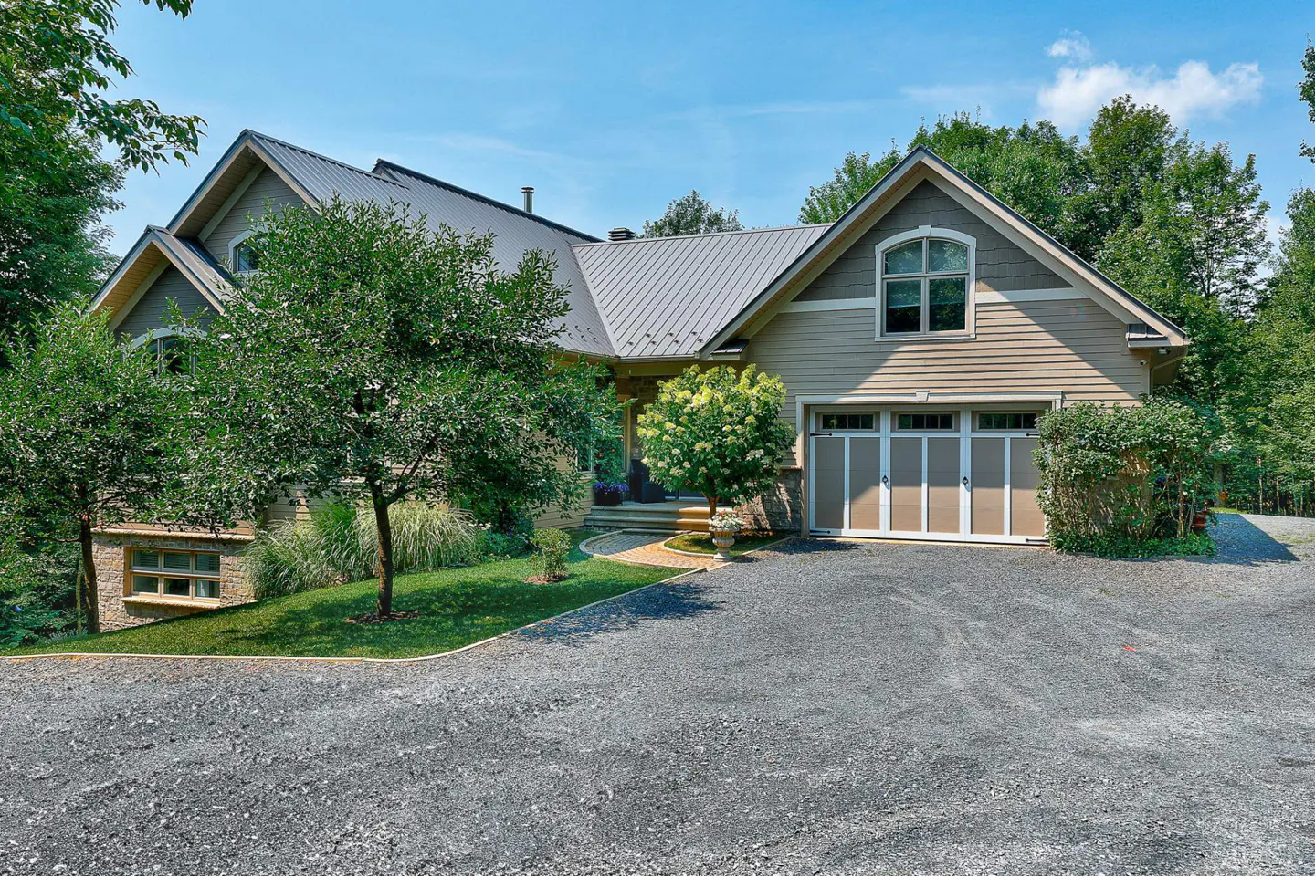 A tan house with a gray metal roof and a two-car garage is surrounded by green trees and a gravel driveway.