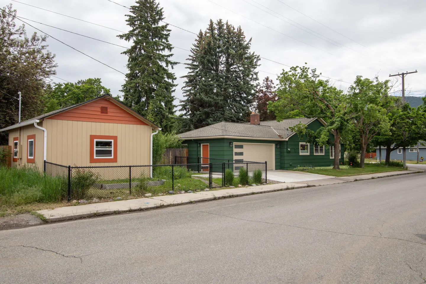 A tan house with orange trim next to a green house with a garage, set along a quiet street with tall pine trees.
