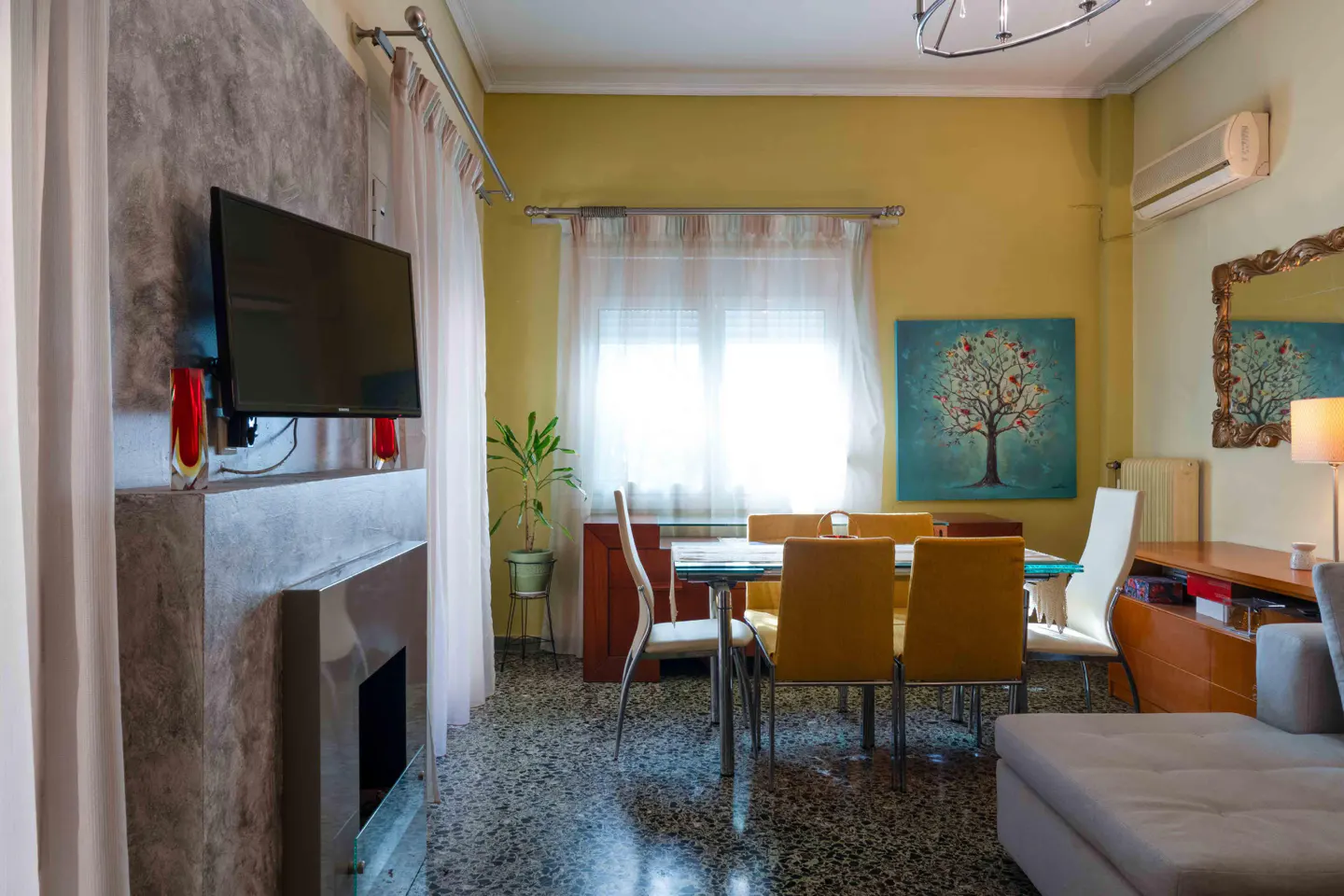 Living room with a TV above a modern fireplace, a glass dining table with yellow chairs, and a painting of a tree on the wall.