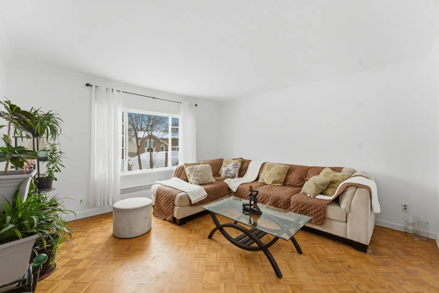 Living room with parquet floors, a brown sectional sofa with pillows, and a glass coffee table. Plants near a window with a snowy outdoor view.