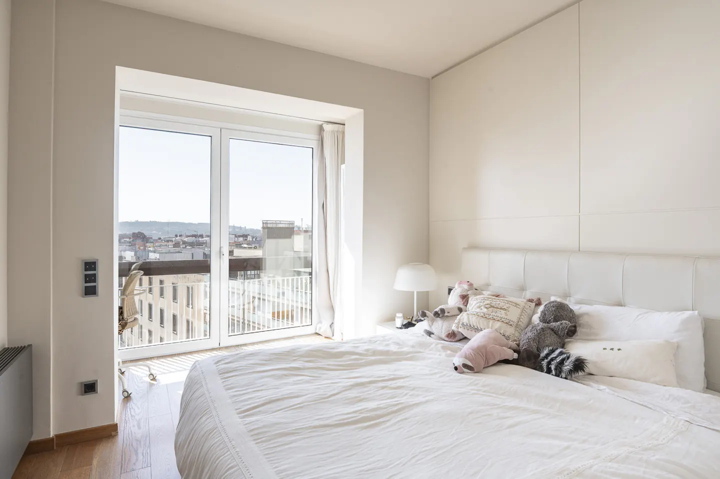 Bright bedroom with a white bed, pillows, and stuffed animals. A large window overlooks a city skyline.