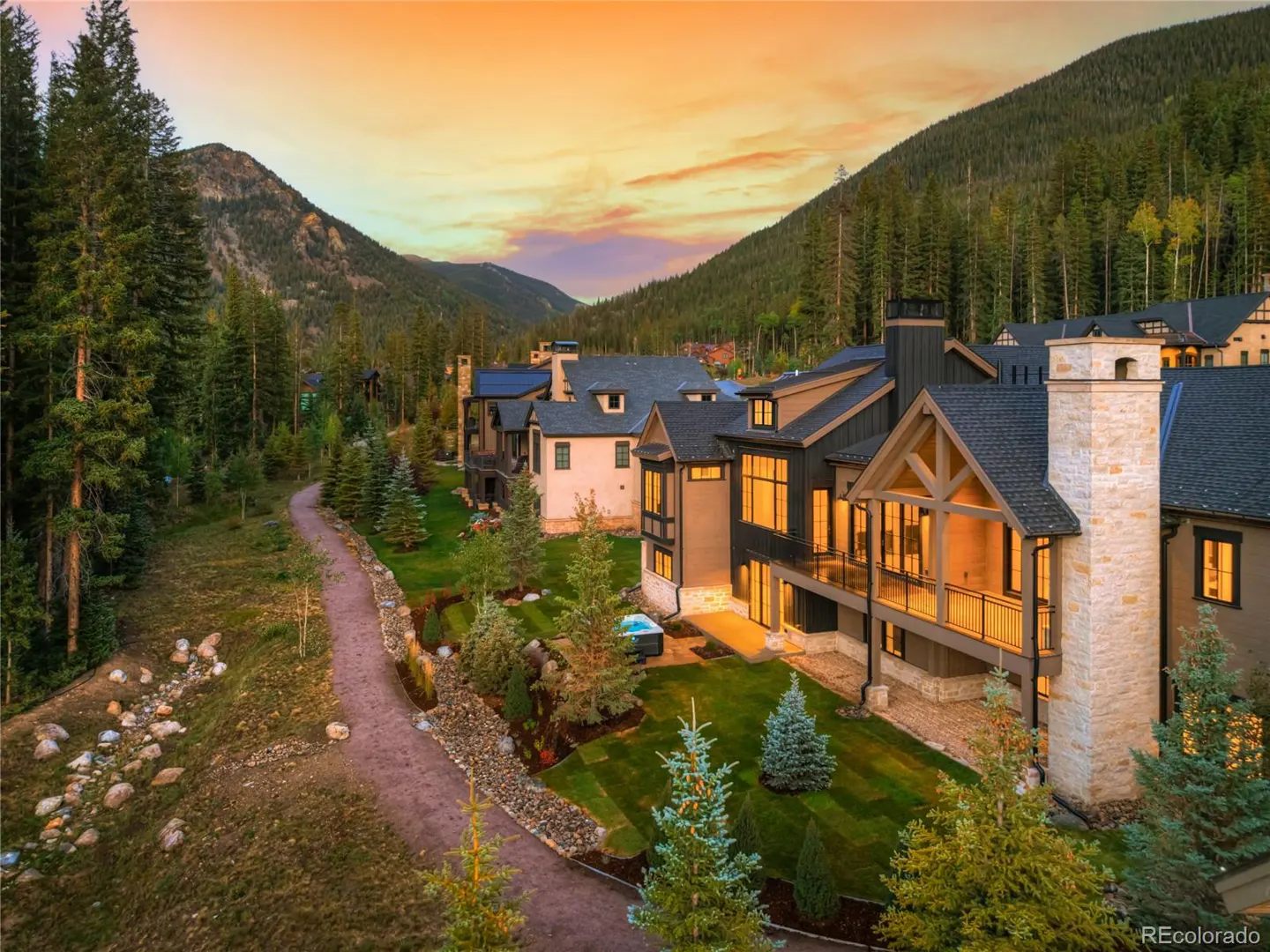 Luxury homes nestled in a mountain valley at sunset. Dark roofs and stone chimneys stand out against the green trees.