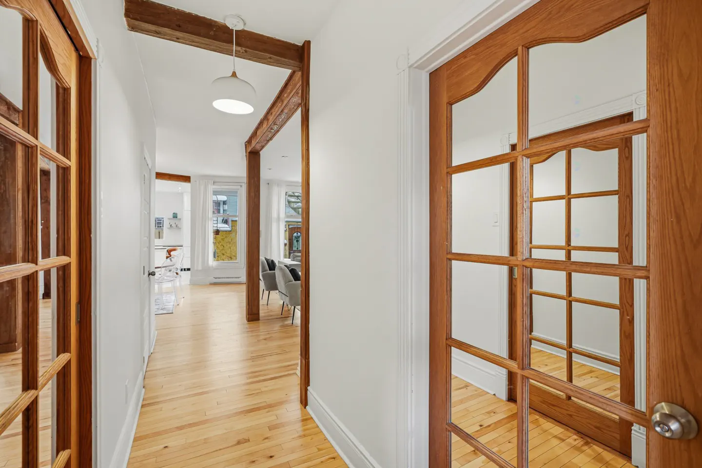 Hallway with light wood floors, white walls, and exposed beams. A glass-paneled door is on the right, and a living room is visible in the background.