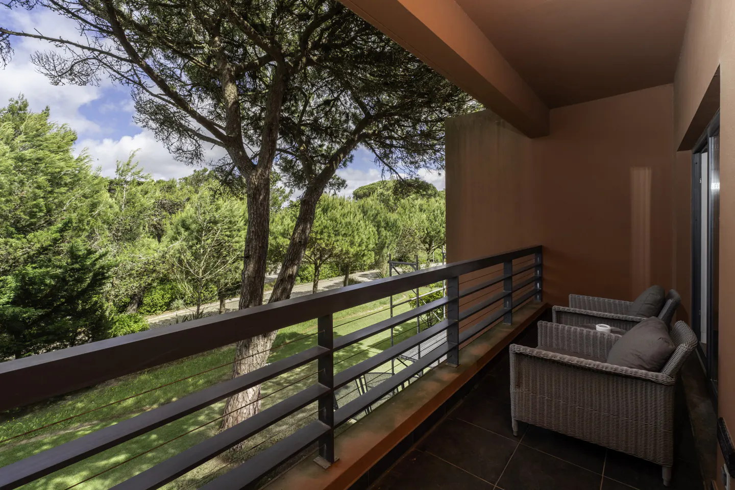 Balcony with two wicker chairs and a view of green trees and blue sky. The balcony has a brown railing and a red floor.