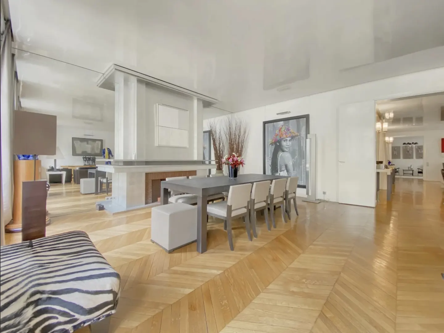 Bright, modern dining room with a gray table, beige chairs, and herringbone wood floors. A large portrait hangs on the wall.