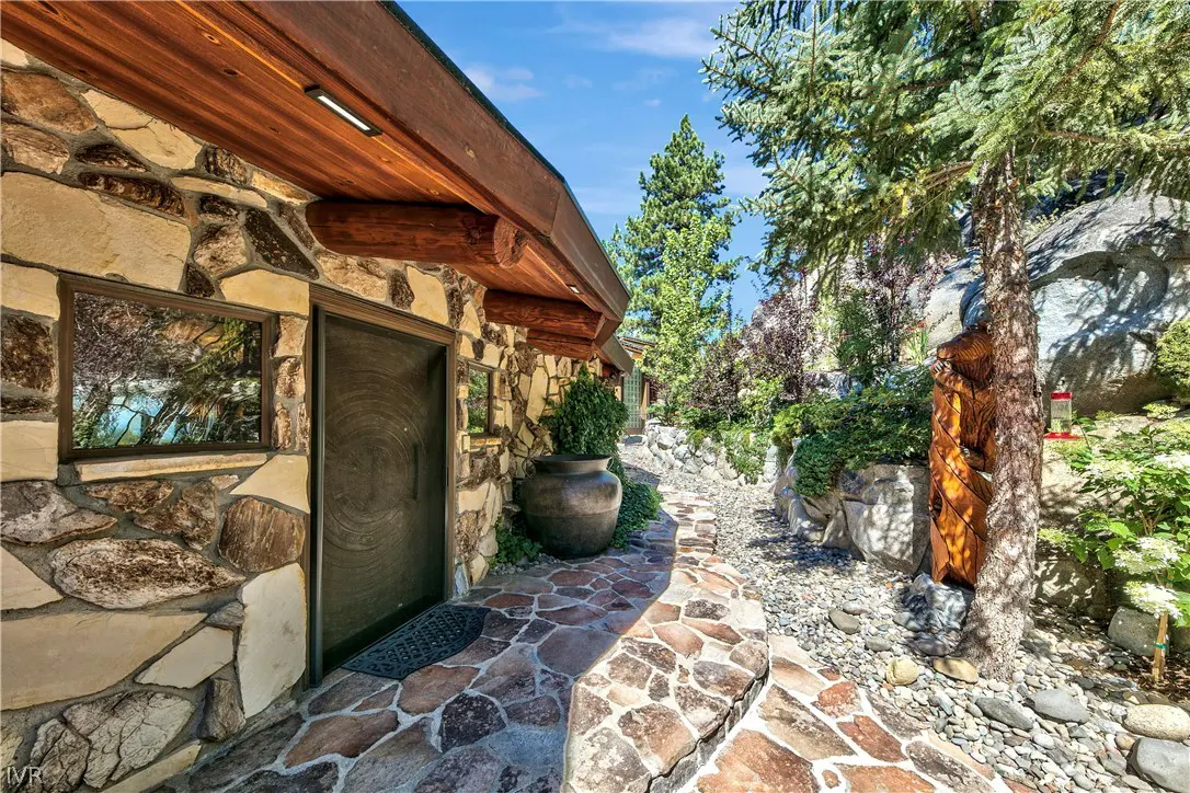 Stone house exterior with a dark door, flagstone path, large decorative pot, and surrounding trees under a blue sky.