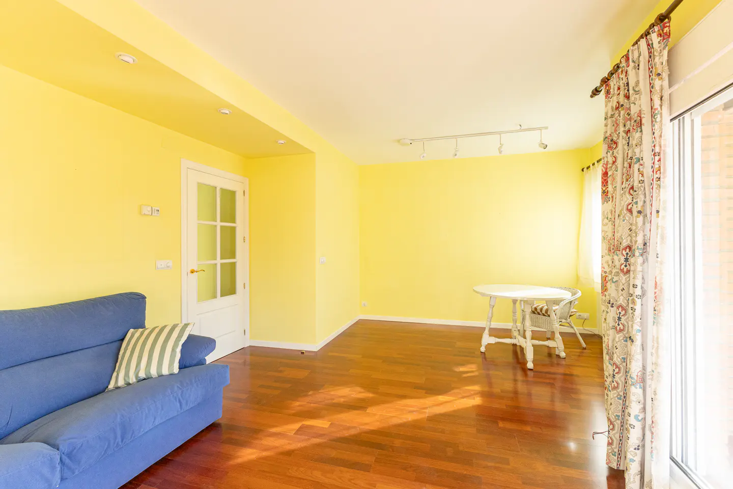 Bright yellow living room with hardwood floors, a blue sofa, and a white table near a window with floral curtains.