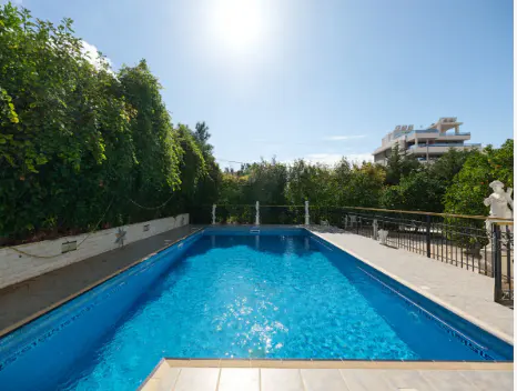Outdoor pool with clear blue water, surrounded by lush green trees and a building in the background under a sunny sky.