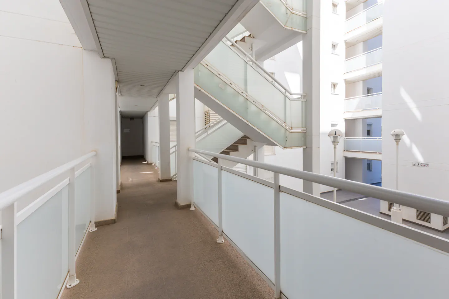 Hallway with white walls, gray railings, and a concrete floor. A staircase with glass panels is visible.