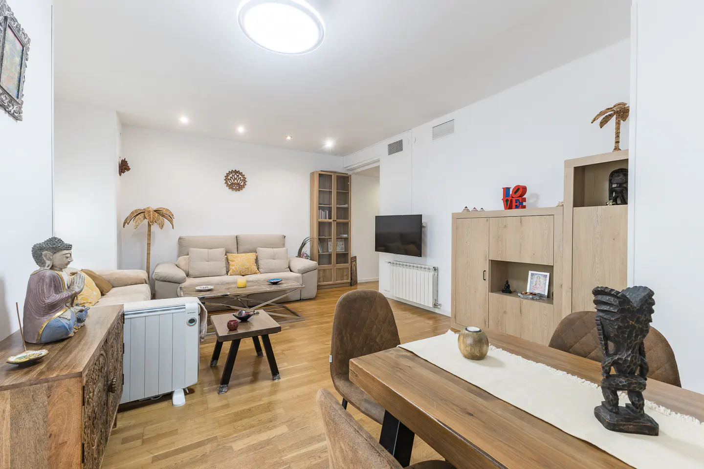 Bright living room with hardwood floors, beige sofas, wooden furniture, and white walls. A dining table with chairs is in the foreground.