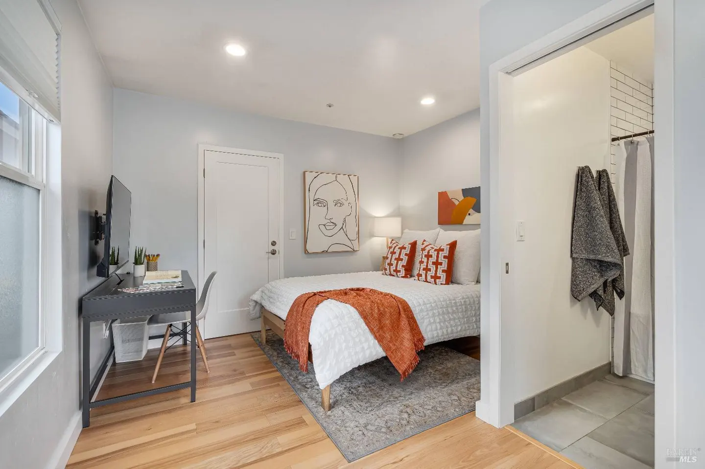 Bedroom with a queen bed, orange accent pillows, a desk, and an attached bathroom with a shower and hanging towels.