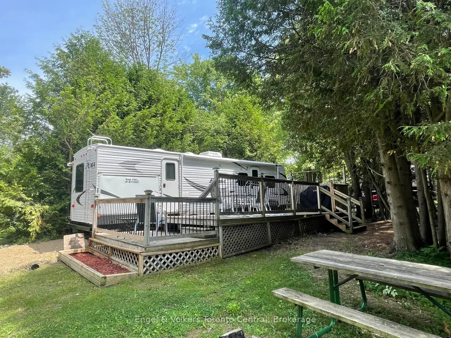 Exterior view of a white RV with a wooden deck, surrounded by green trees and grass. A picnic table sits in the foreground.