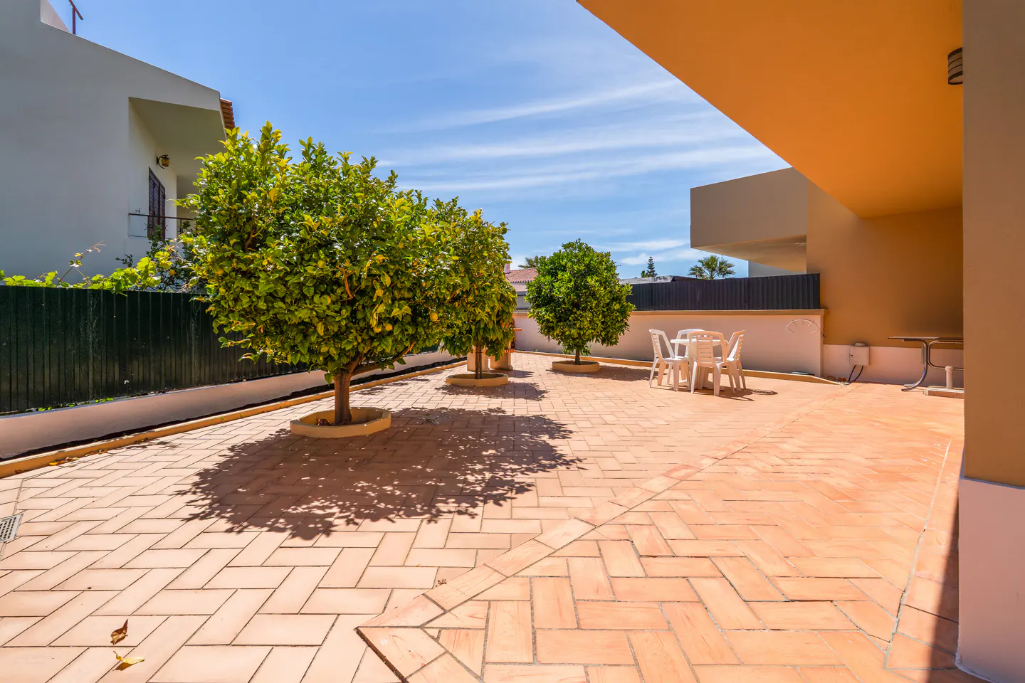Outdoor patio with brick pavers, green trees, and white table and chairs. Blue sky in the background.