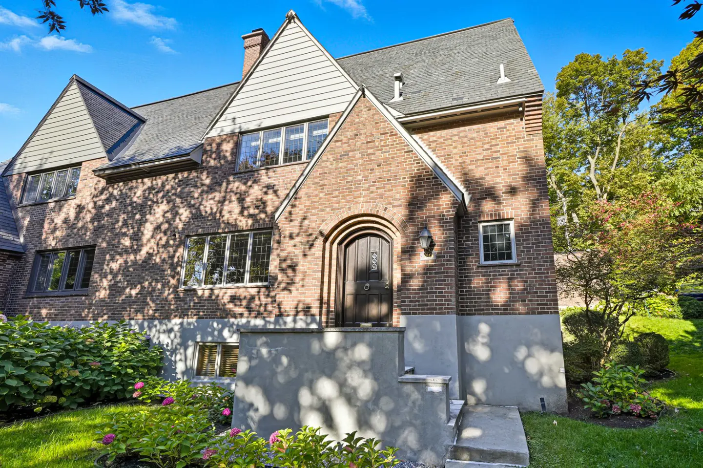 Exterior of a brick Tudor-style home with a dark wood arched front door, gray roof, and green landscaping.