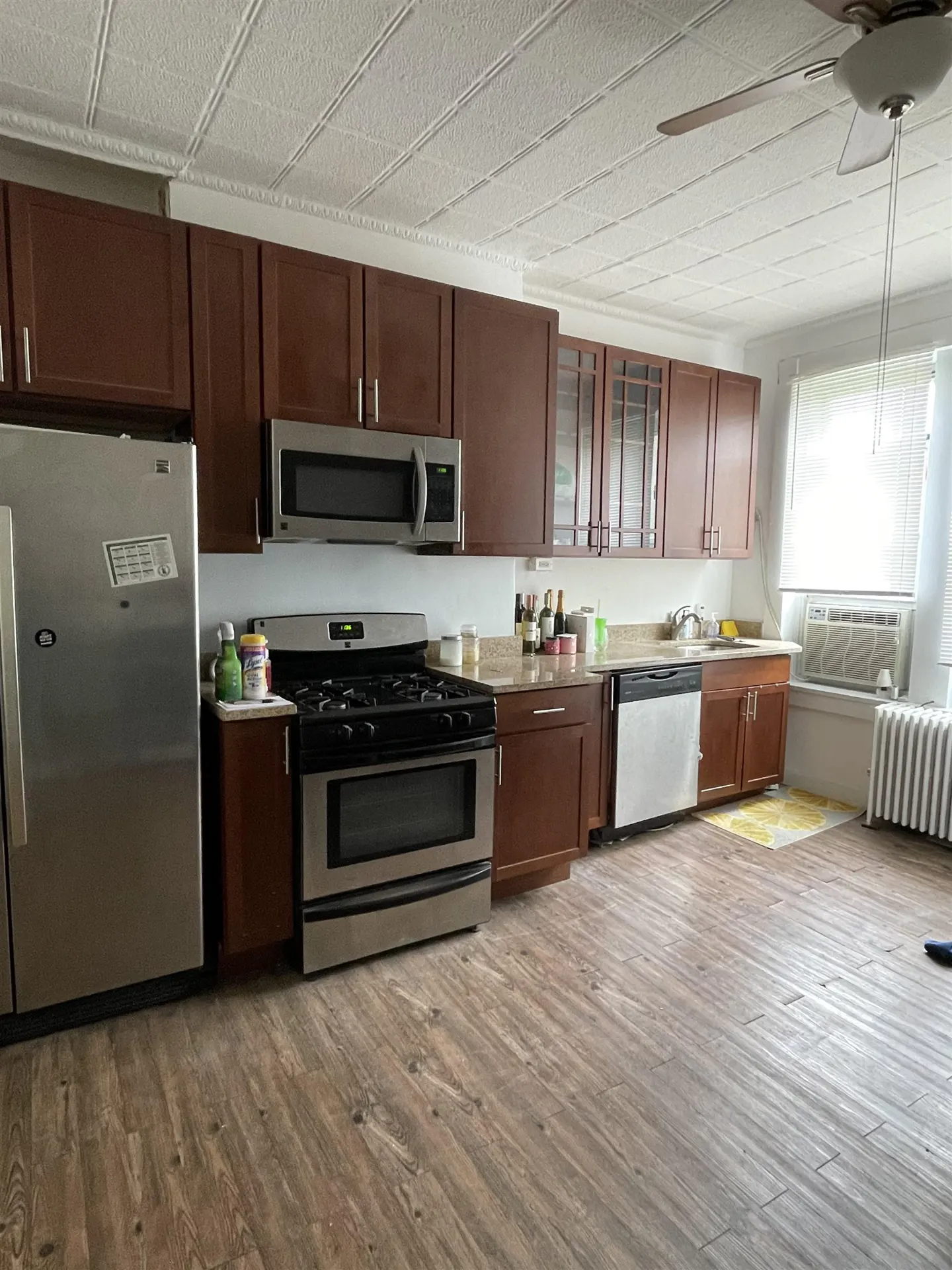 A kitchen with brown cabinets, stainless steel appliances, and wood-look flooring. A window with an AC unit is visible.