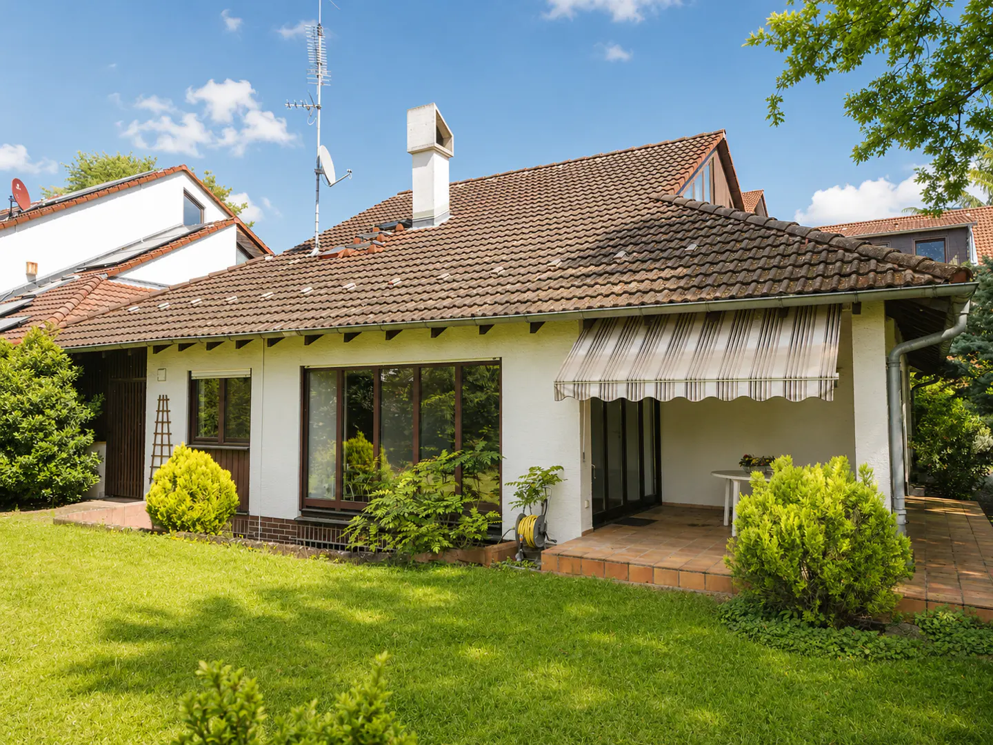A single-story white house with a brown tile roof, green lawn, and blue sky. A striped awning covers the patio.