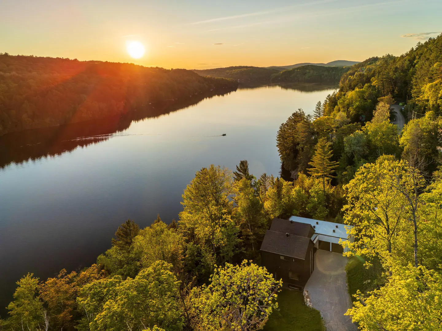 Aerial view of a dark house nestled in lush green trees beside a calm lake at sunset with a golden sky.