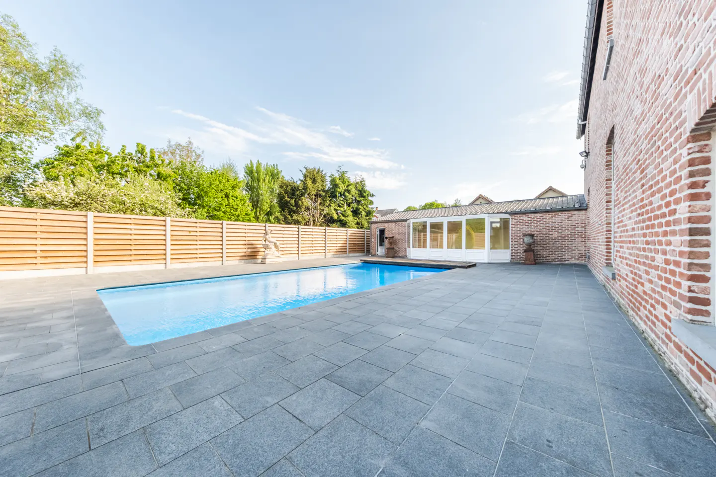 A backyard with a blue swimming pool, gray stone patio, and a red brick building. A wooden fence lines the yard, with green trees in the background.