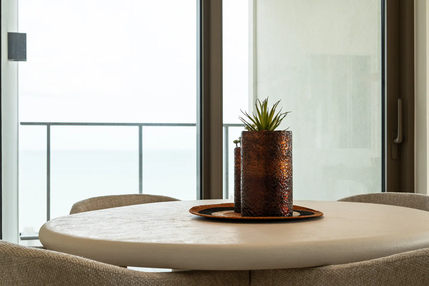 A round white dining table with a textured bronze vase and plant centerpiece, set before a window with a balcony view.