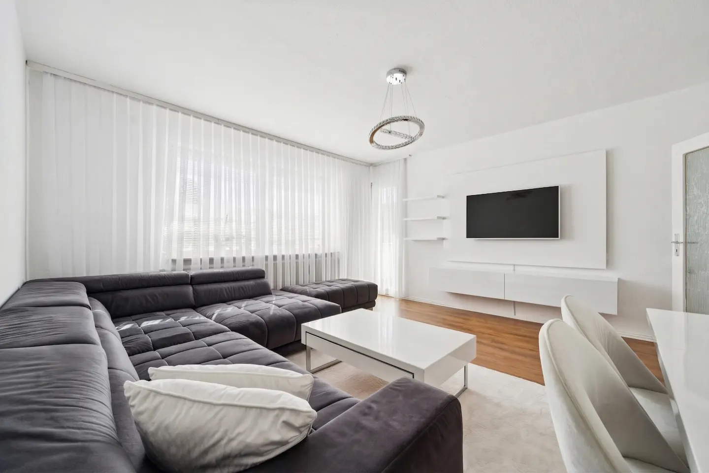 Bright living room with a gray sectional sofa, white pillows, and a white coffee table. A TV is mounted on the wall with white shelves and cabinets.