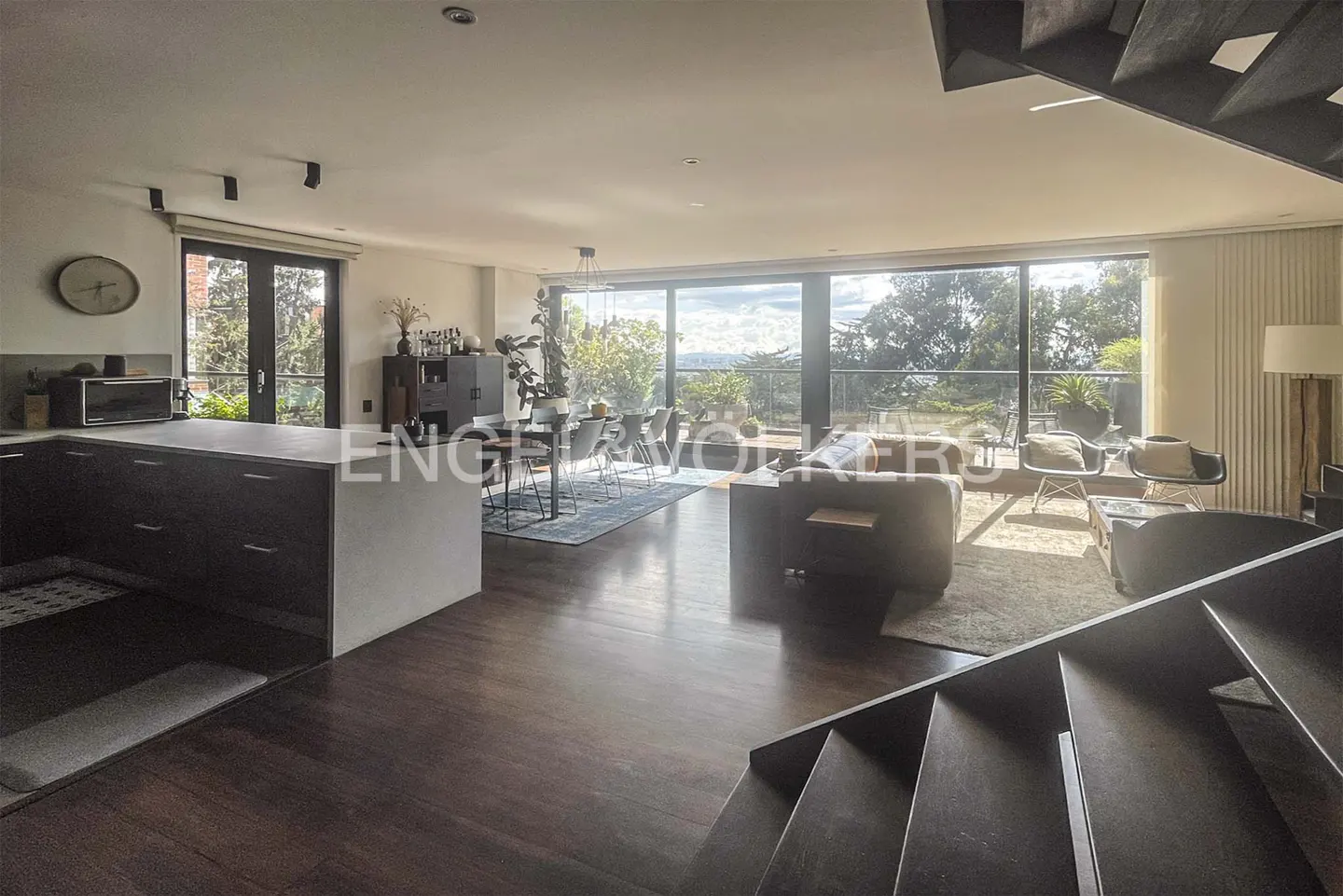 Open-concept living space with dark wood floors, kitchen island, dining table, and living room with large windows overlooking greenery.