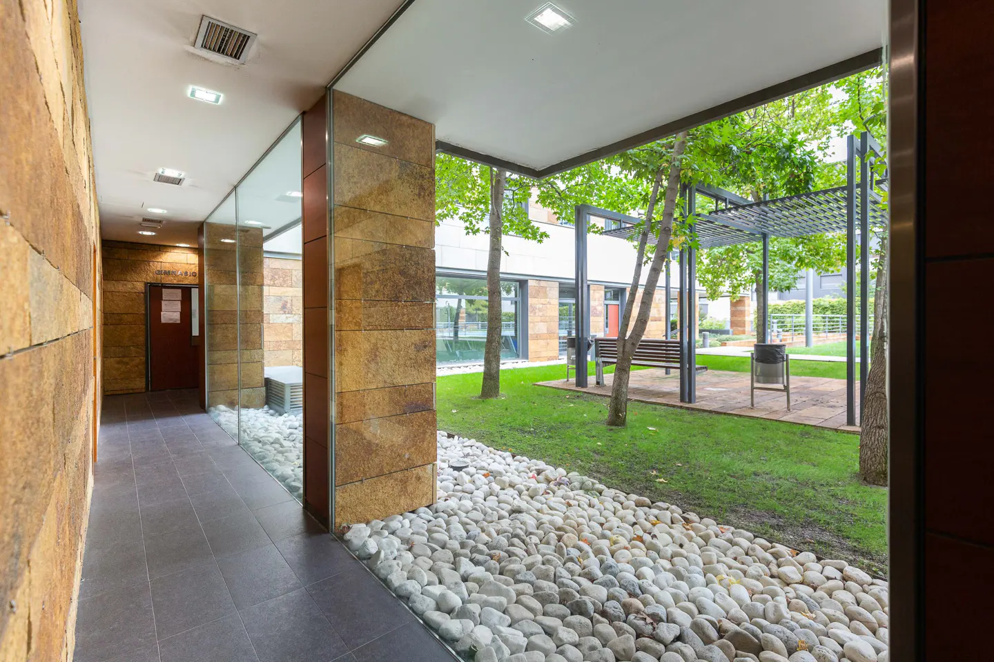 Hallway with stone walls and tile floor leads to a courtyard with green grass, trees, and a bench. White pebbles line the base of the wall.