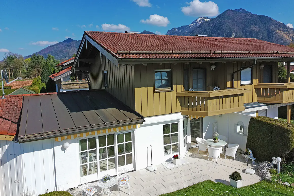 Two-story house with brown roof and balconies, white patio with table and chairs, and mountains in the background.