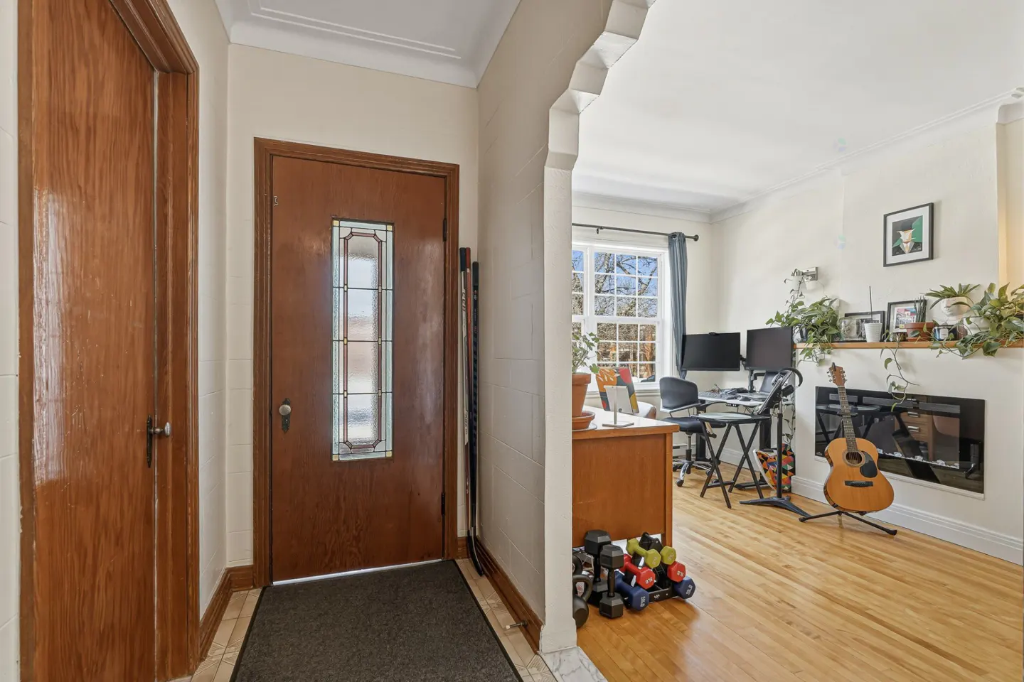 A home's entryway with a brown front door with a glass window, and a view into a living room with a desk, fireplace, and guitar.