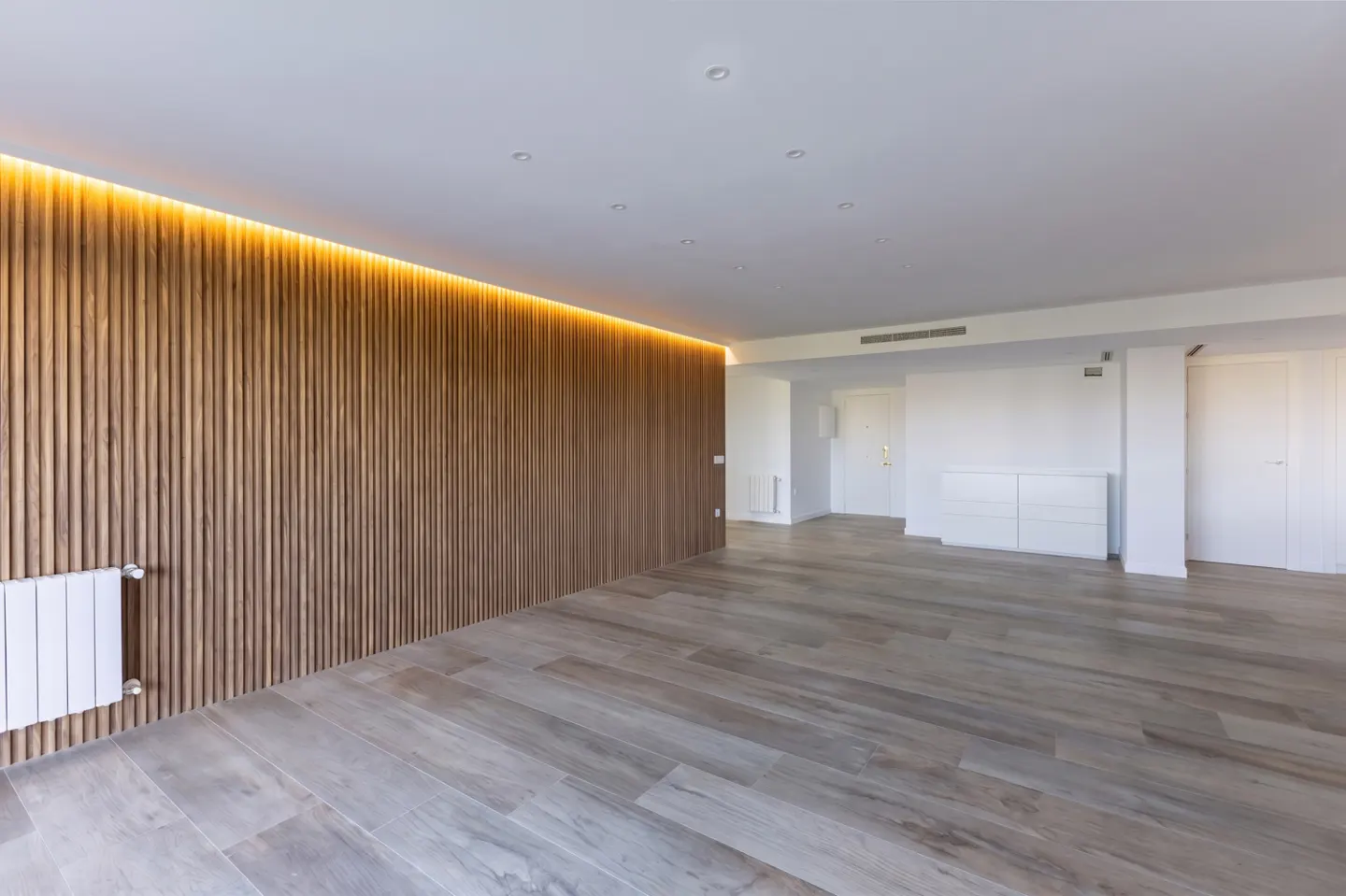 An empty room with wood-look tile floors, a wood slat wall with warm lighting, and white walls.