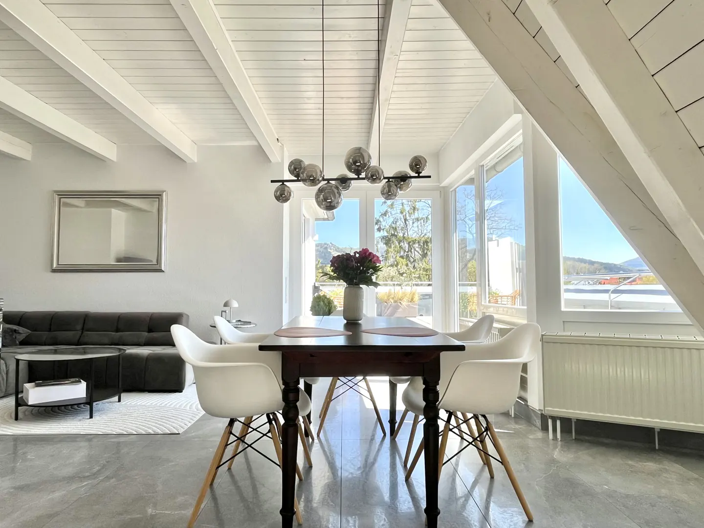 Bright, modern dining room with a dark wood table, white chairs, and a unique bubble chandelier. Large windows offer a scenic view.