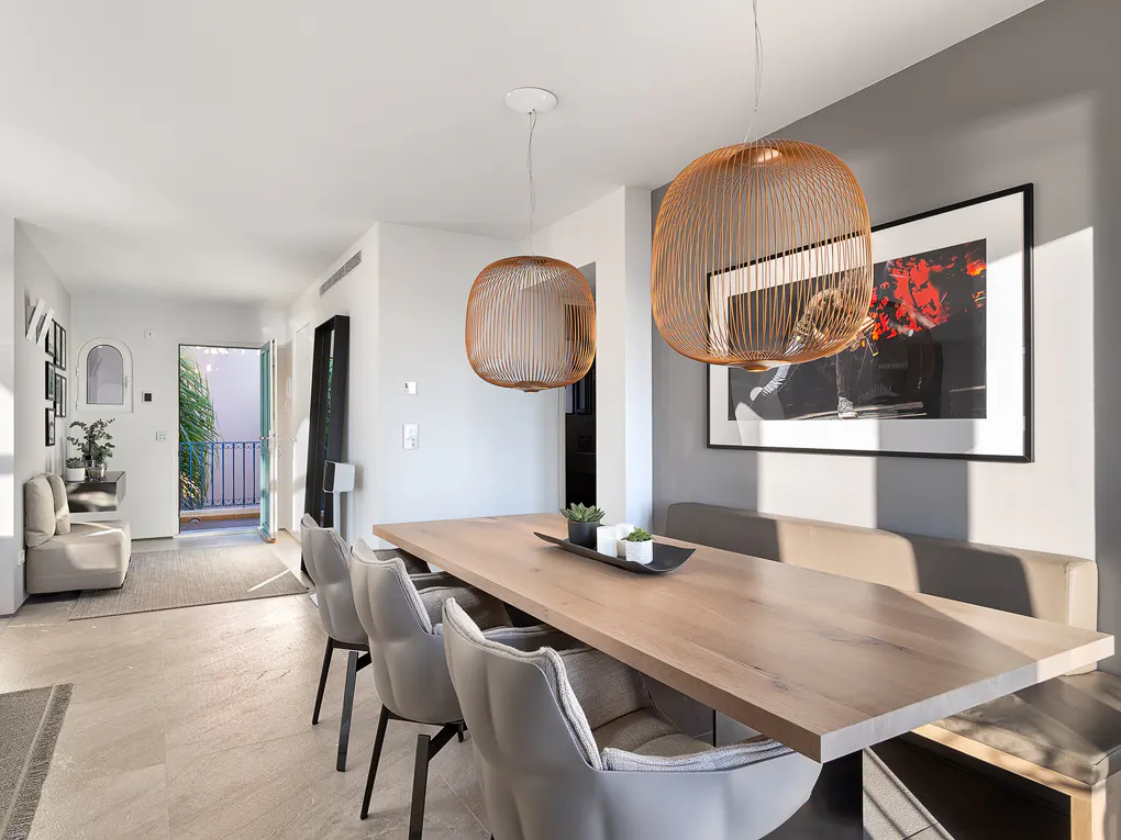 Bright dining room with a long wooden table, grey chairs, and two copper pendant lights. A large framed art piece hangs on the grey wall.