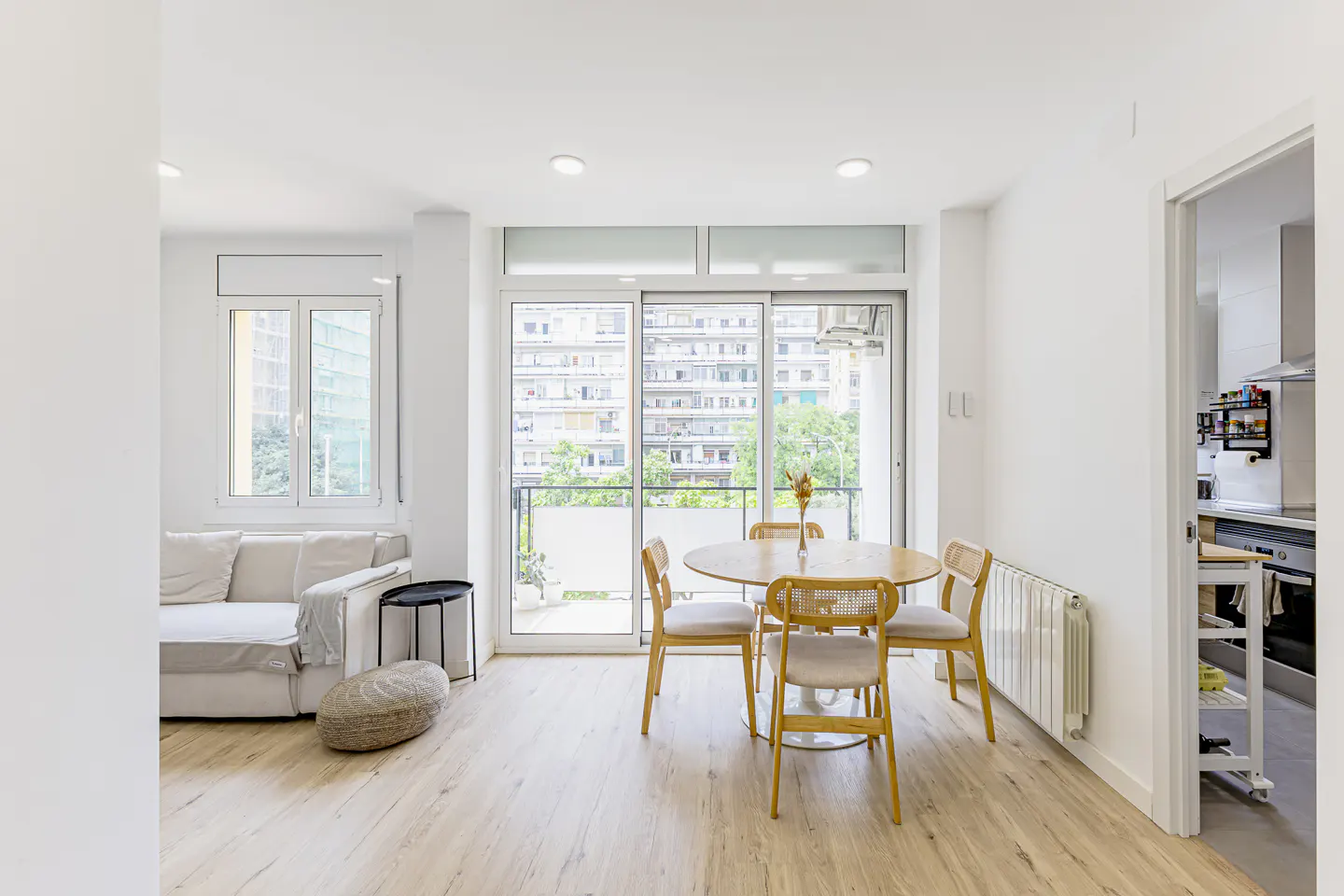 Bright living room with light wood floors, white sofa, and a round dining table with four chairs by a balcony door.