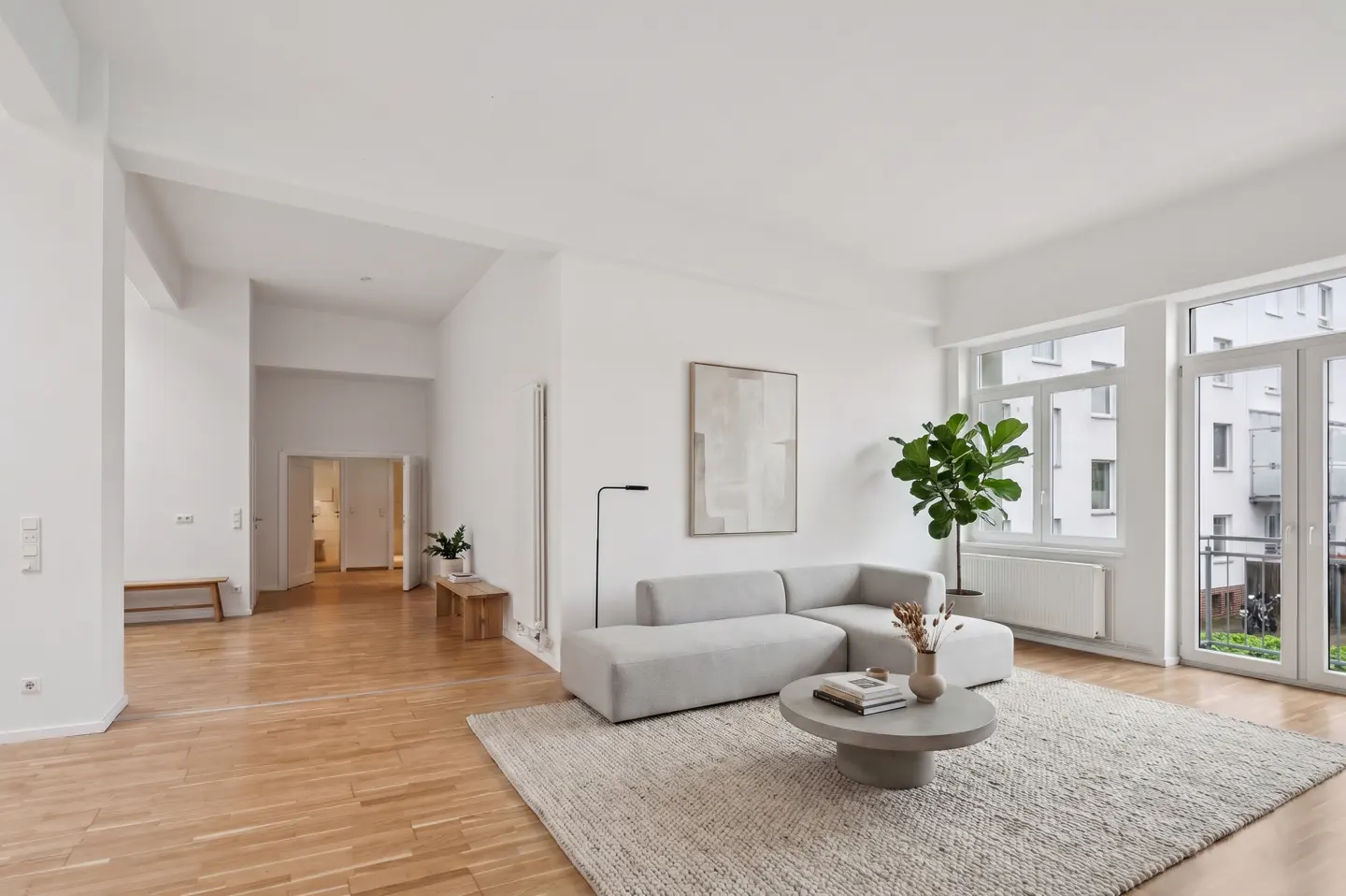 Bright, modern living room with light wood floors, white walls, and a gray sectional sofa on a textured rug. Large windows offer a view of an apartment building.