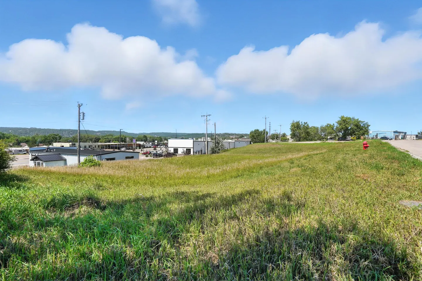 A grassy lot with commercial buildings in the background under a blue, cloudy sky. A red fire hydrant is on the right.