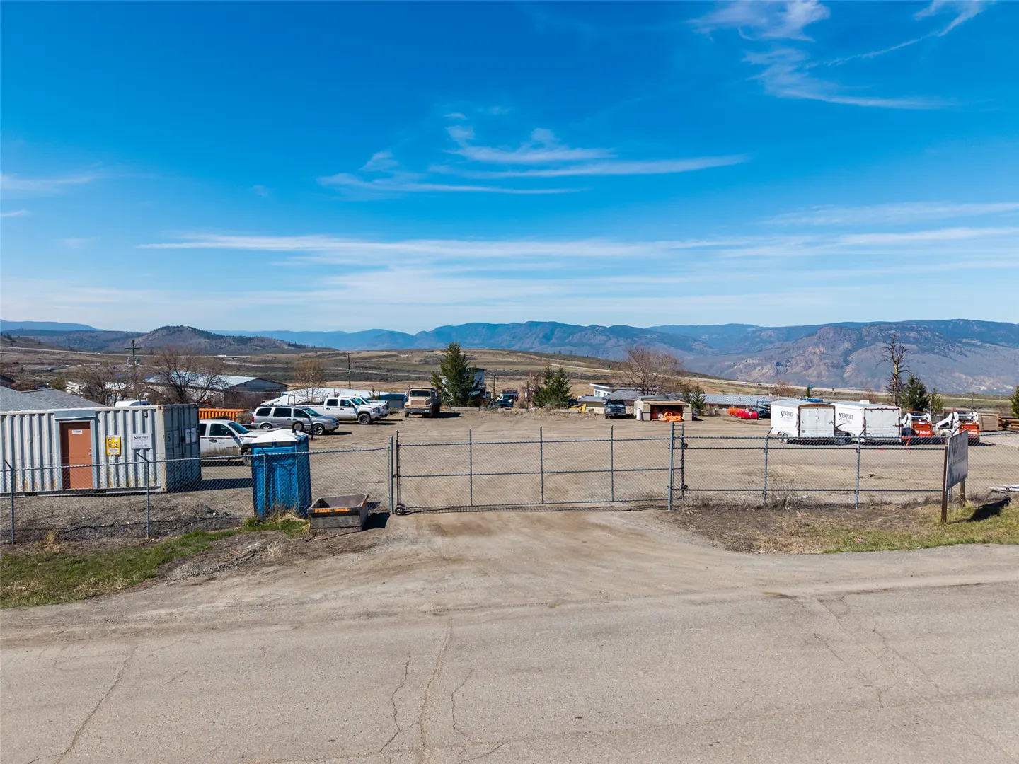 Gravel lot with a metal gate, storage containers, trucks, and equipment under a blue sky with mountains in the distance.