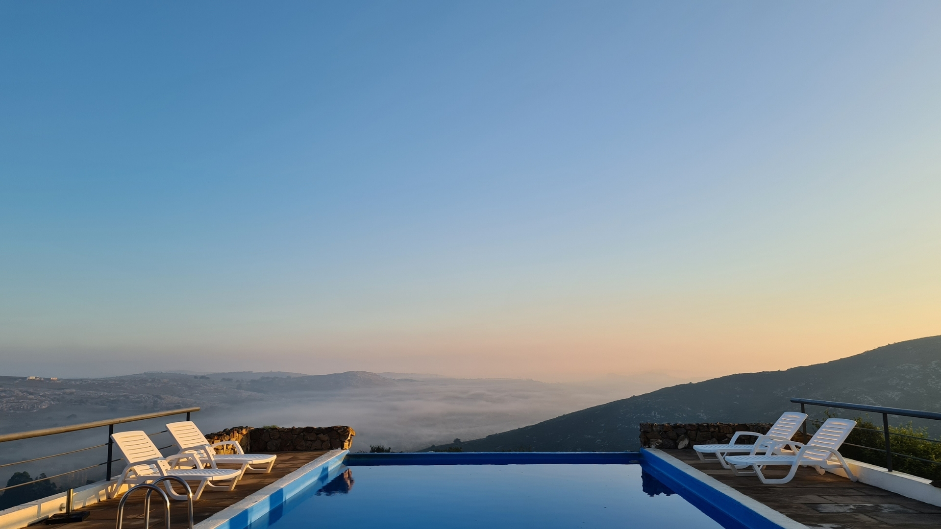 Infinity pool on a deck with white lounge chairs overlooking a misty mountain valley at sunrise. [AI-generated text]