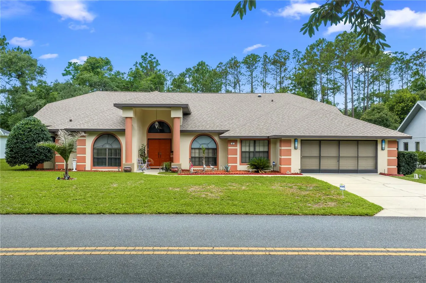 Beige single-story house with a brown roof, arched windows, and a two-car garage, set against a backdrop of green trees and a blue sky.